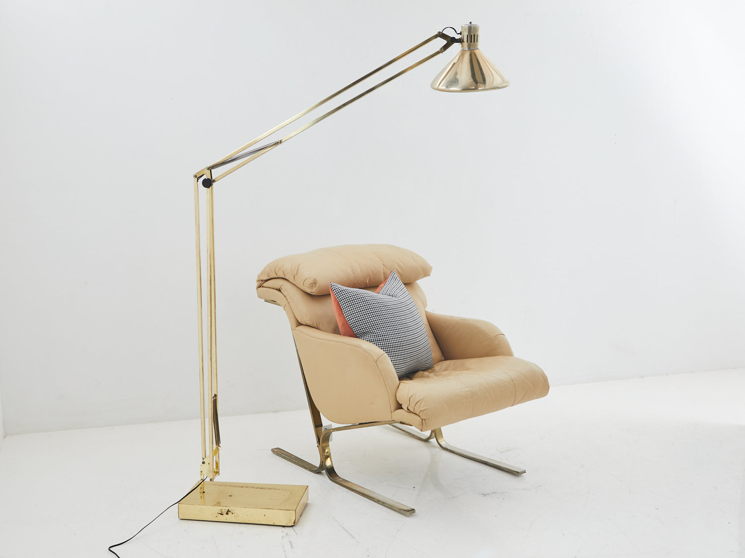 Brass adjustable floor lamp to the left of a tan lounge chair with brass feet