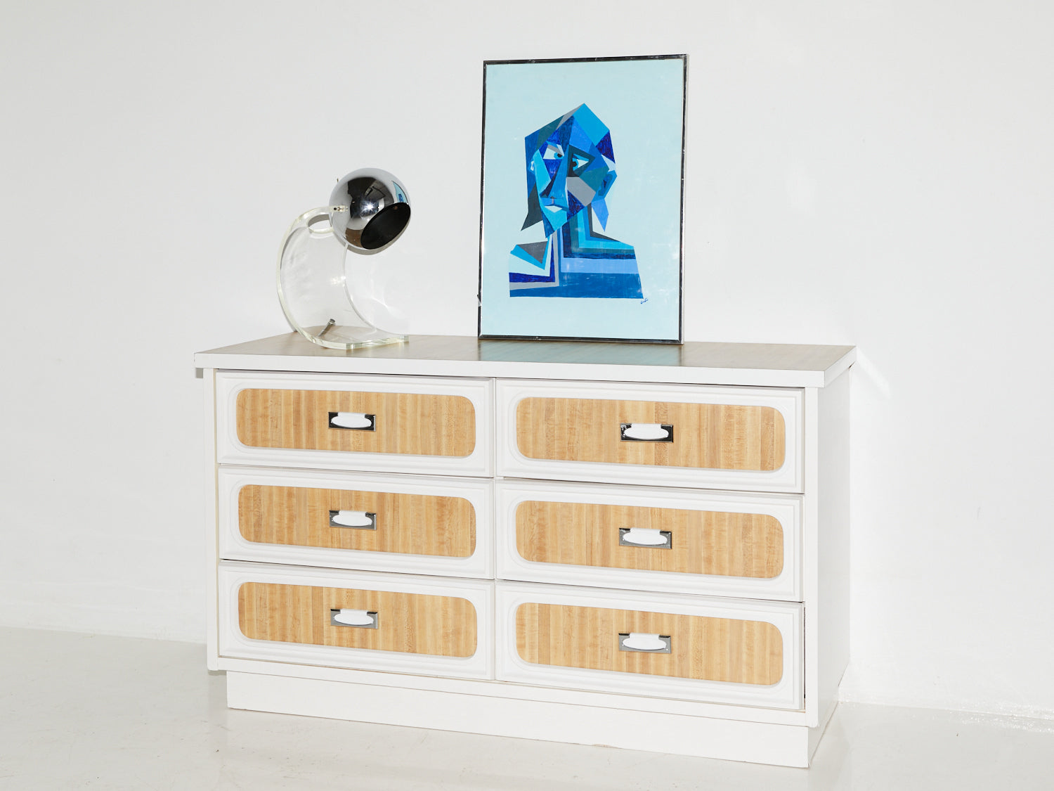 Faux-wood formica dresser with 6 drawers with a Sonneman style lamp and acrylic painting on top